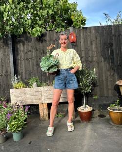 Woman in a yellow shirt and denim shorts holding a large succulent in her garden.