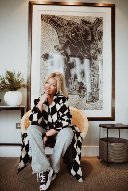 Woman sitting in a chair wearing a black and white coat and jeans, in front of a large framed print.