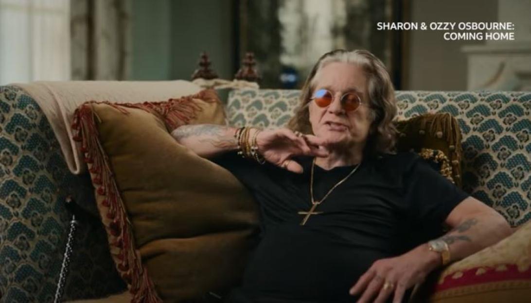 Ozzy Osbourne sitting on a sofa in a black shirt and red-tinted glasses.