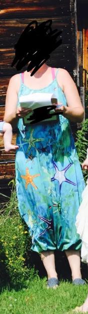 An officiant in a blue sundress with starfish patterns and visible pink bra straps, holding papers, standing in grass.