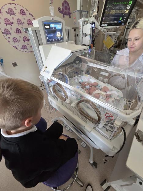 A newborn in an incubator, watched by an older sibling and a woman.