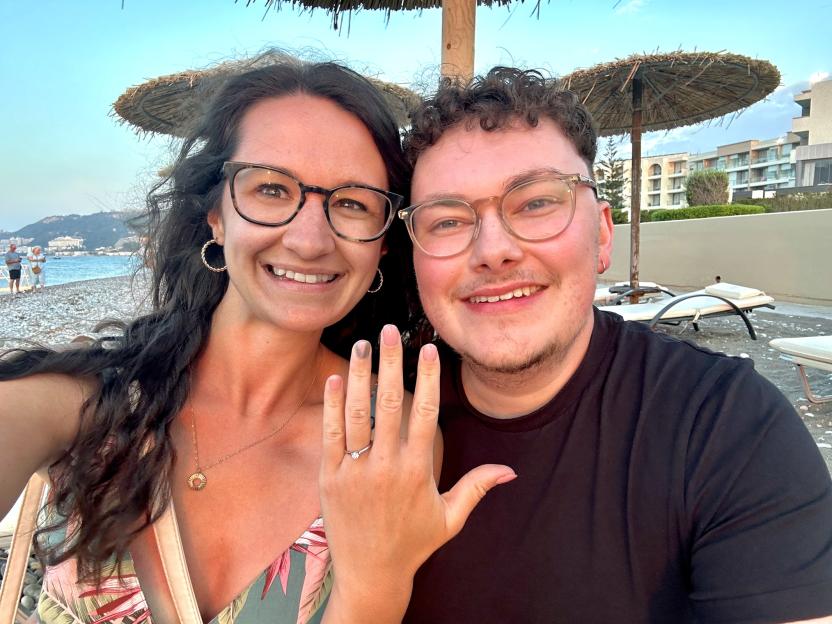 Georgia and Laurence posing on a beach, with Georgia showing her engagement ring.