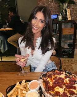 Woman holding a glass of wine at a restaurant table with pizza and fries.