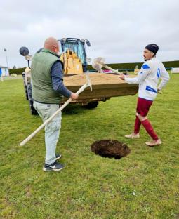 Two men filling a sinkhole on a soccer field with a digger.