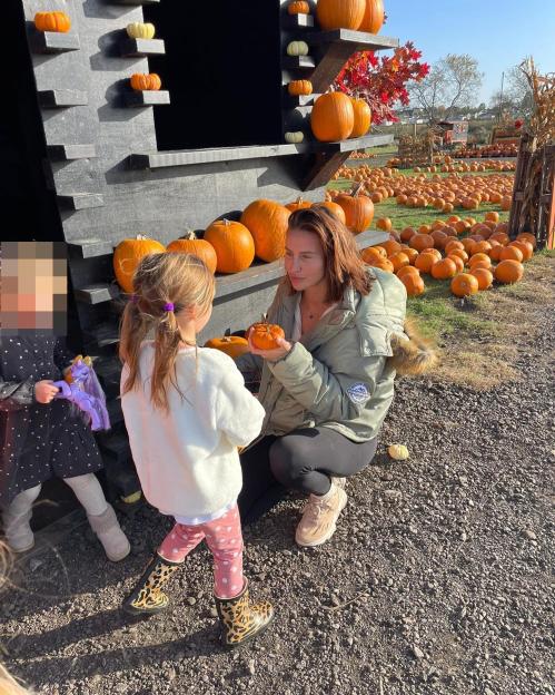 Ferne McCann kneels in a pumpkin patch, showing a small pumpkin to her daughter Sunday, who has her back to the camera.
