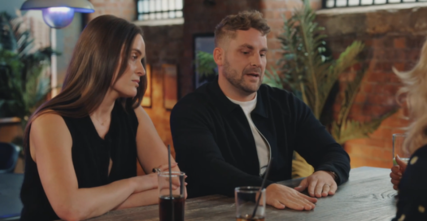 A man and a woman sitting at a table with drinks, looking at someone off-camera.