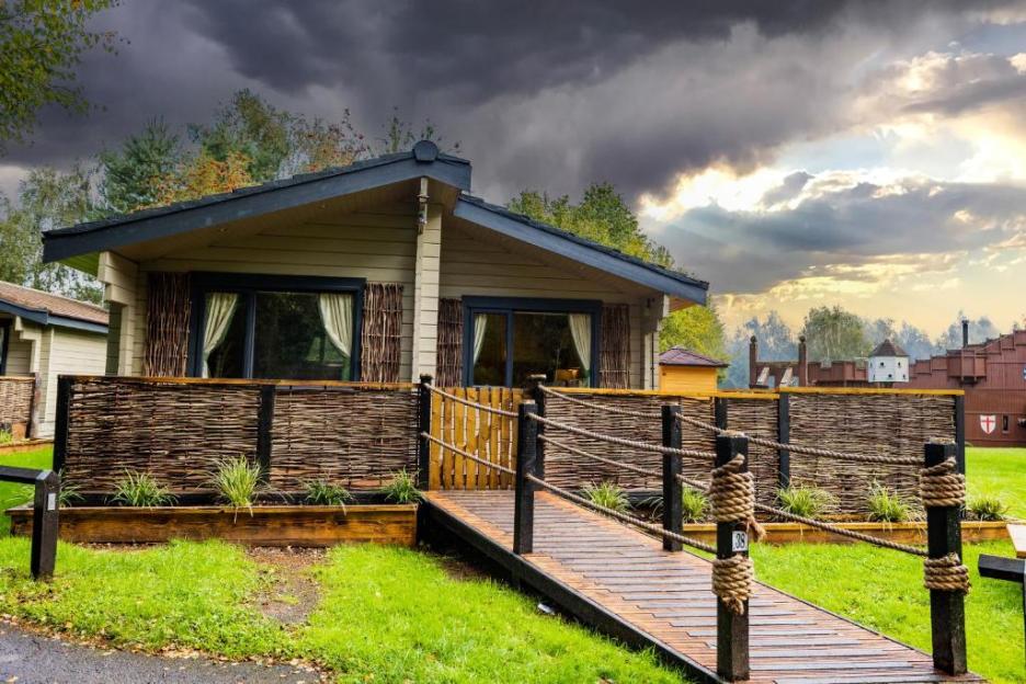 Cabin with a wooden walkway and fence, near a medieval-style building.