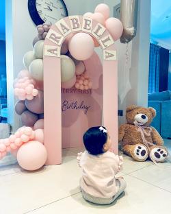 Baby girl sitting in front of Arabella's first birthday balloon arch and teddy bear.