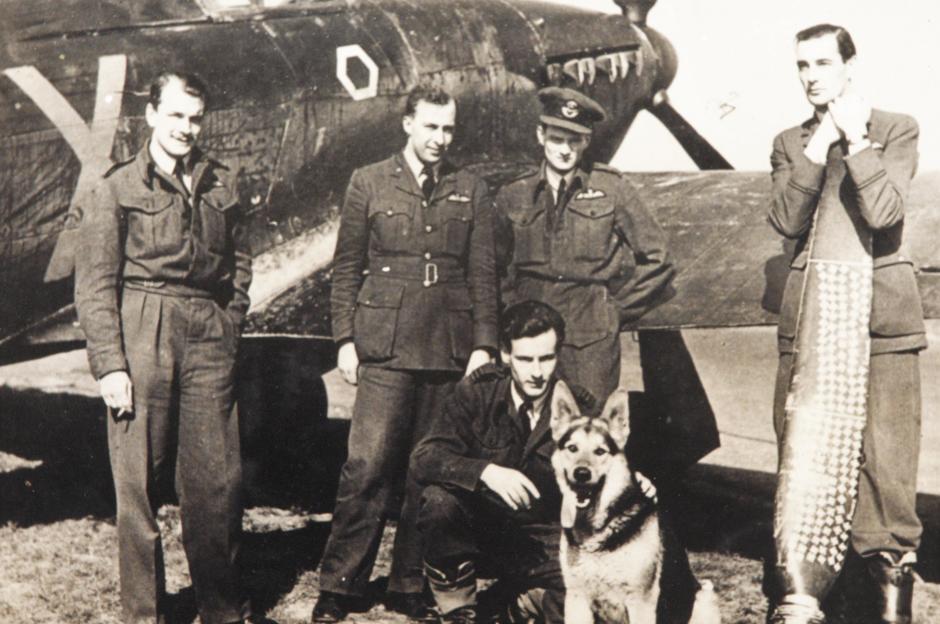 Black and white photo of five men in military uniforms and a German Shepherd dog in front of an airplane.