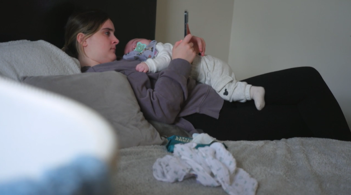 A woman lying in bed holding a baby while looking at a phone.