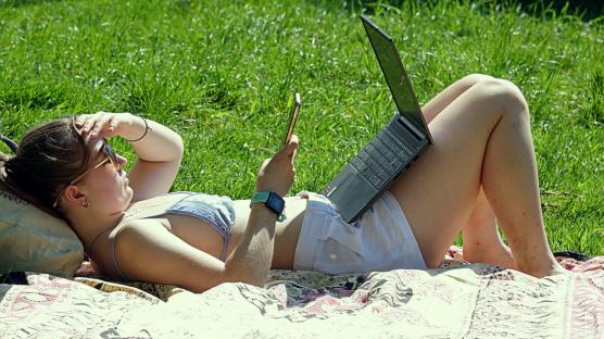 Woman sunbathing on grass using laptop and phone.
