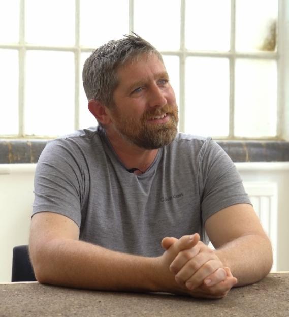 A man sits at a table, smiling and looking off to the side.