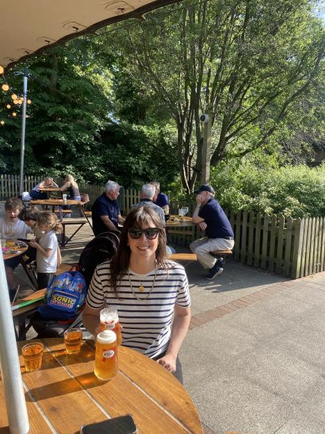 A woman holding two glasses of beer smiles at the camera while sitting at an outdoor restaurant.