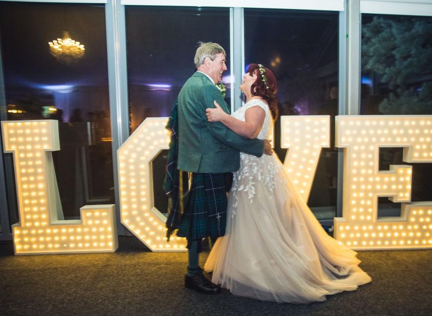 Patrick Howard with his wife Laura at their wedding, standing in front of large illuminated letters spelling "LOVE".
