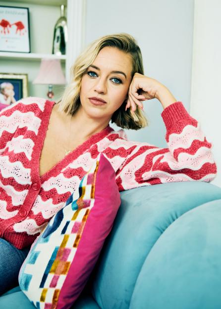 Kelsey Parker resting her head on her hand on a blue couch, wearing a pink and red cardigan.