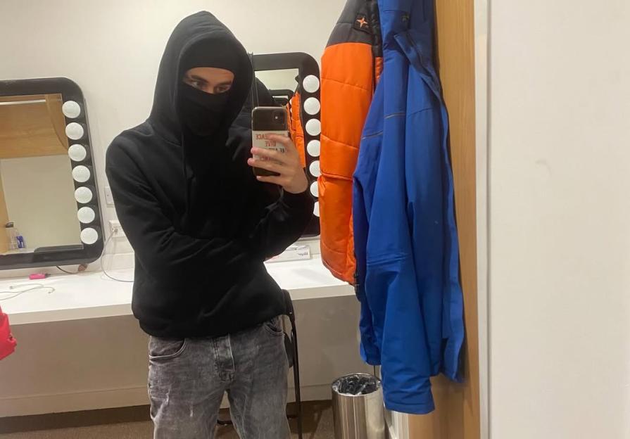 A person wearing a black hoodie and balaclava takes a selfie in a dressing room.