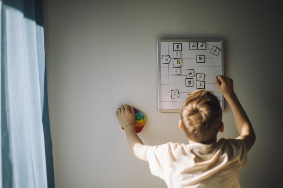 A boy with autism holding a rainbow Pop It toy while interacting with a visual aid whiteboard.