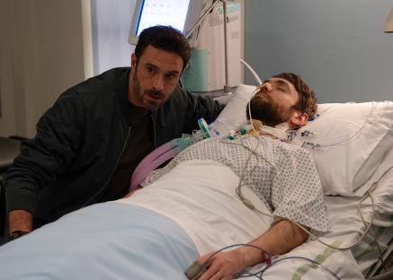A man looking concerned sits beside a man lying in a hospital bed.