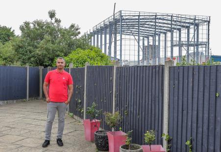 Man in red shirt standing in his garden next to a fence, with a large metal warehouse frame under construction visible in the background.
