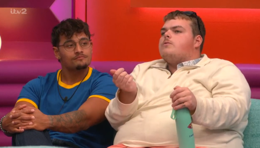 Two men sitting on a couch, one speaking and holding a bottle while the other listens.