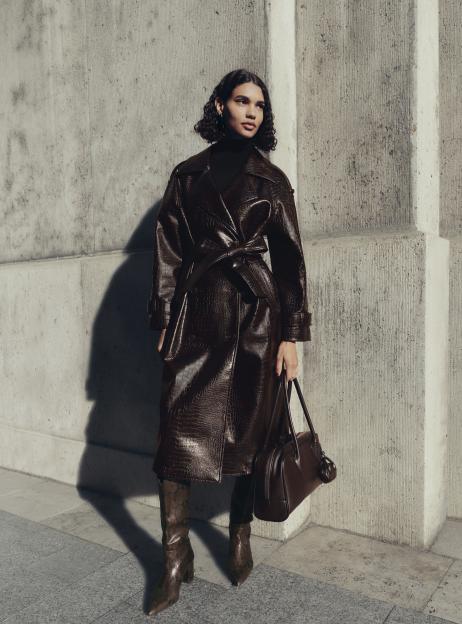A model in the M&S Autumn Winter 2025 collection wearing a brown crocodile-patterned trench coat, dark turtleneck, snakeskin boots, and holding a brown leather bag.