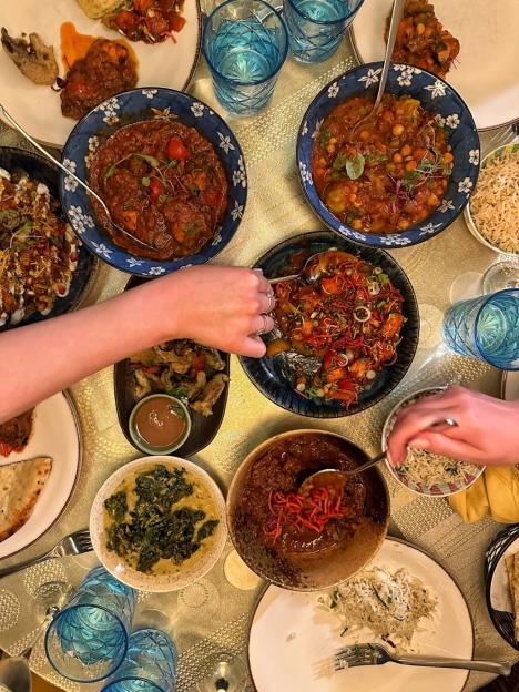 Overhead view of a dinner table laden with various Indian dishes and beverages, with hands serving food.