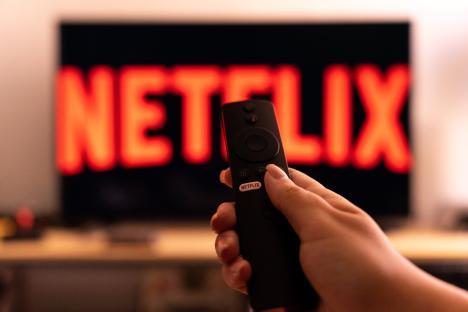 Hand holding a Netflix remote in front of a TV displaying the Netflix logo.
