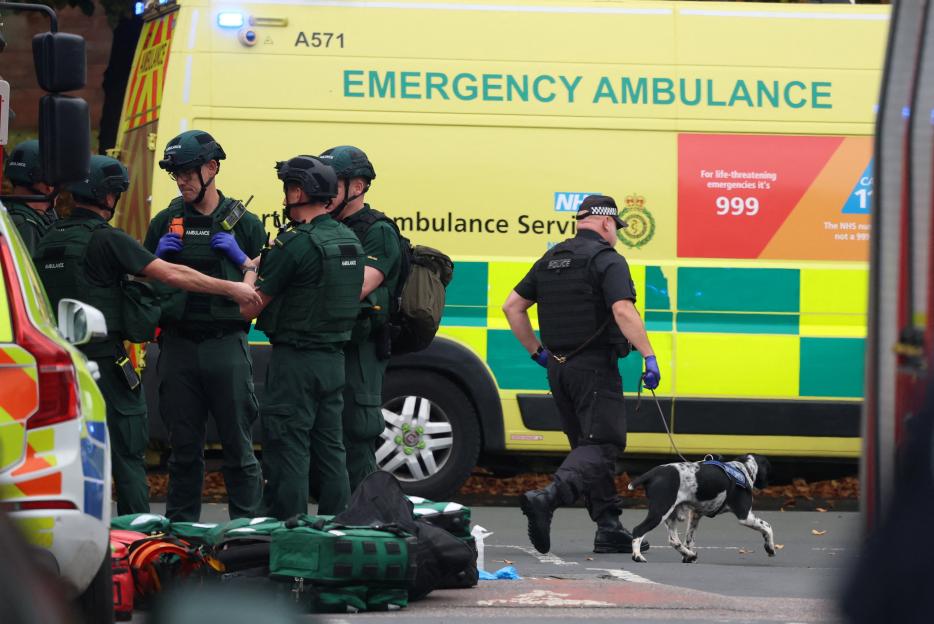 Emergency personnel, including police with a dog and paramedics, work at a scene with an ambulance in the background.